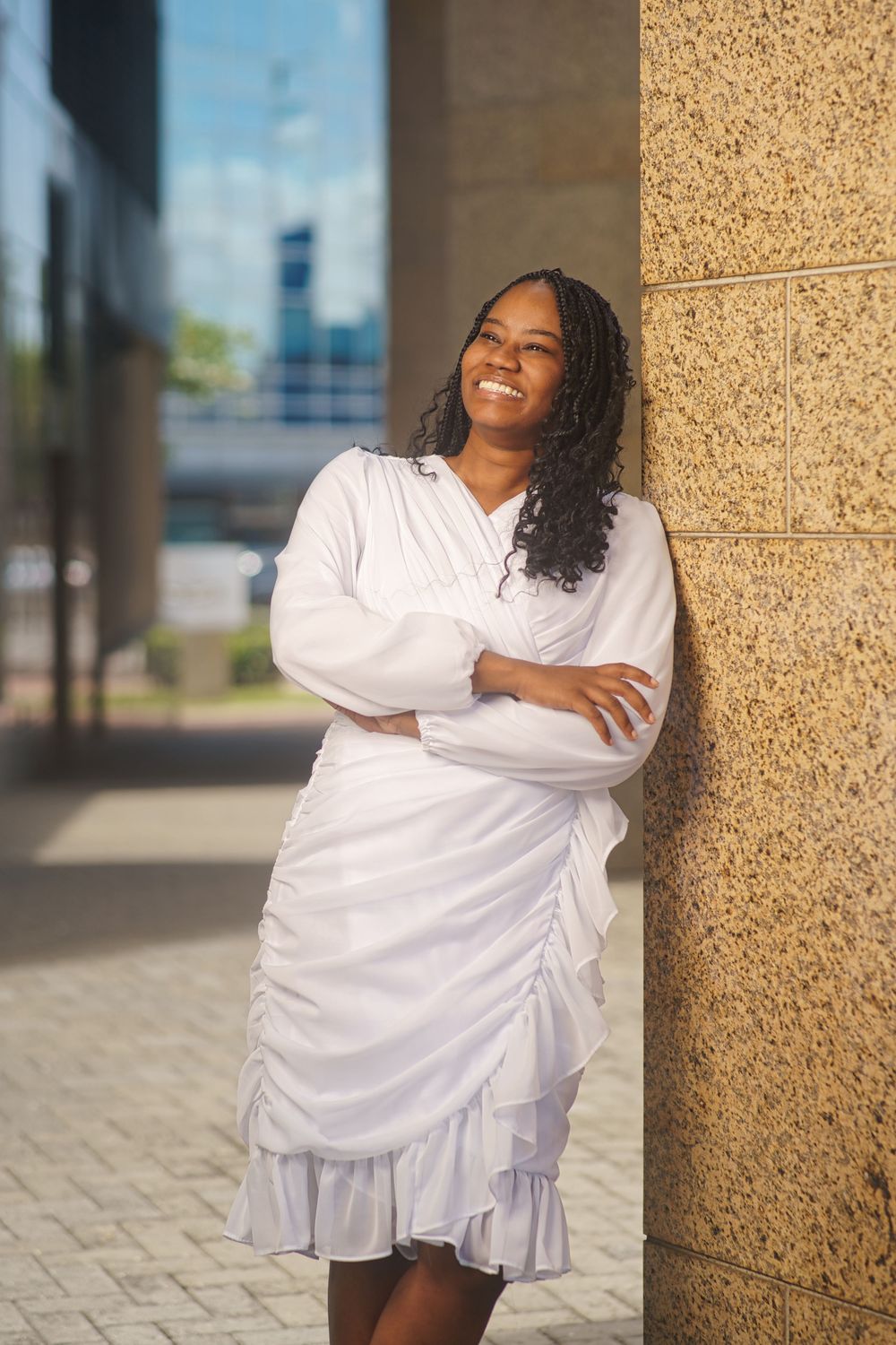 Someone wearing a white ruched dress with ruffled hem poses against a textured wall in an outdoor urban setting.