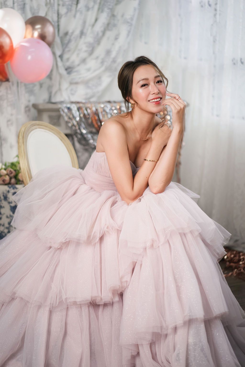 A smiling woman in a pink tulle ball gown poses gracefully with balloons in an elegant decorated room.