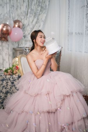 Young woman in pink tulle ball gown holding white birthday cake with crown topper, smiling joyfully.
