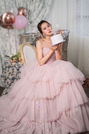 Young Asian woman in pink ball gown holding birthday cake with candles, surrounded by balloons.