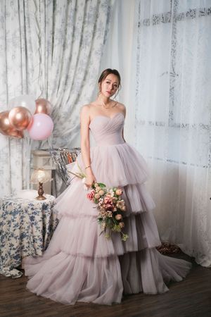 Woman in strapless blush pink tiered tulle ball gown holding floral bouquet near balloons and curtains.