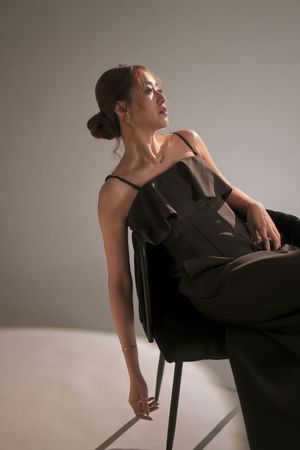 Asian woman in black spaghetti strap ruffled jumpsuit seated on chair, gazing sideways in studio.