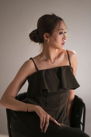 Asian woman in dark olive ruffled cami dress seated on black chair, looking away in soft studio light.