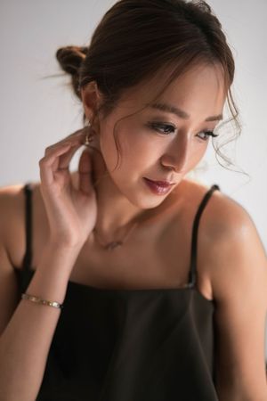 Close-up portrait of Asian woman in black cami top touching ear, wearing bracelet, looking down softly.