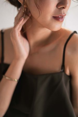 Woman in dark olive slip dress wearing gold bracelet and earring, touching her neck with hand.