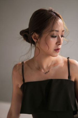 Young Asian woman in black off-shoulder top with updo hairstyle, delicate necklace, and gold earrings poses elegantly.