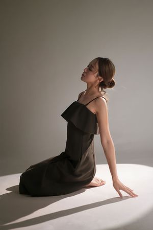 Woman in dark brown spaghetti strap ruffle dress sitting gracefully on floor with head tilted back in studio lighting.
