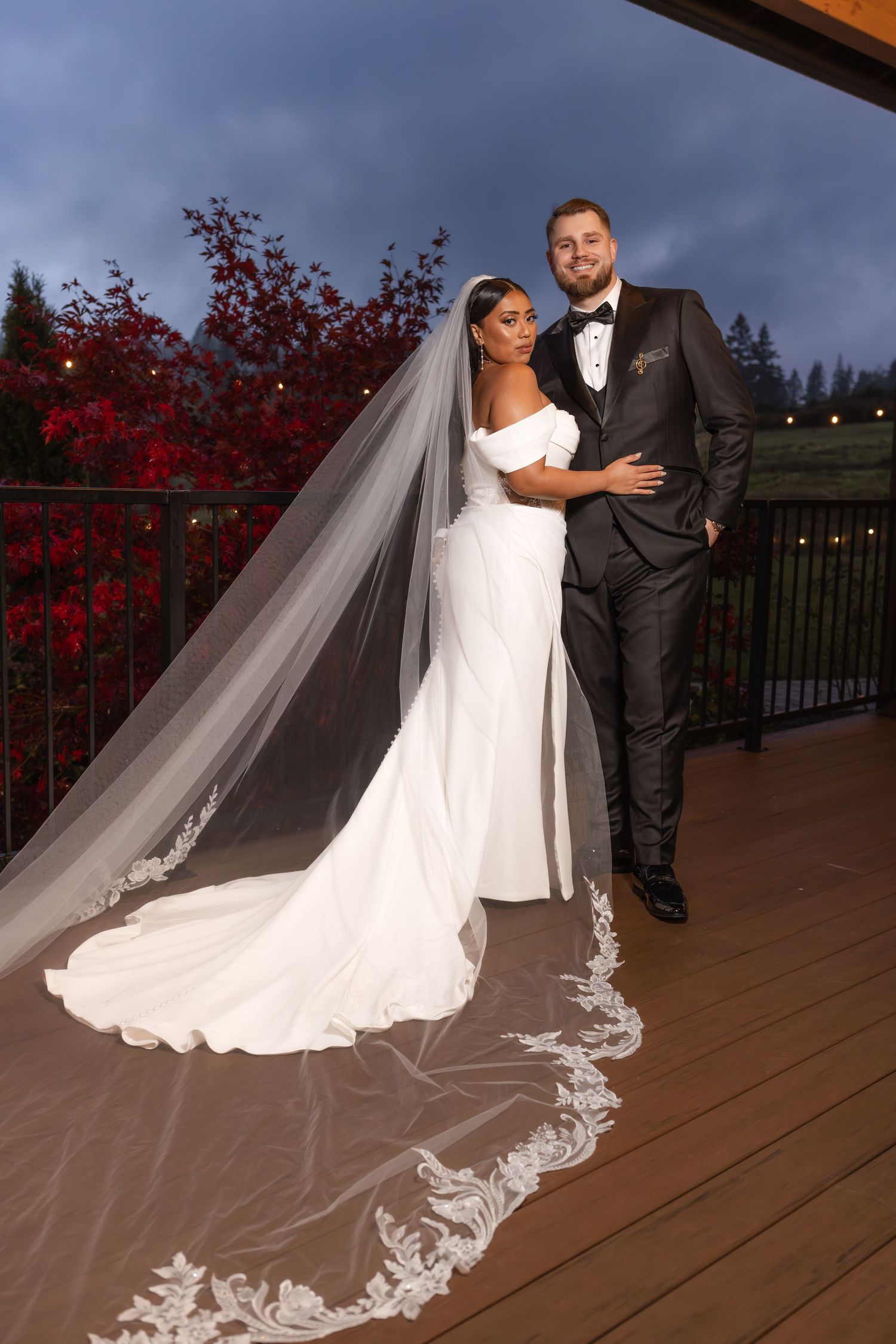 A newlywed couple poses on a wooden deck with vibrant red autumn foliage and dramatic cloudy sky in the background.