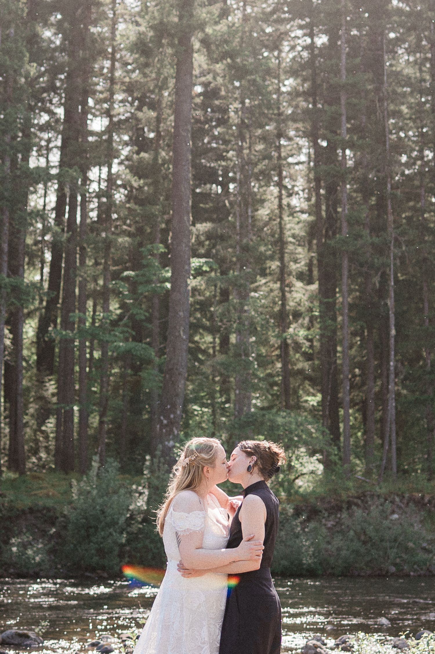 Two brides kiss captured in a sunlit forest setting with pine trees and a small river in the background. A rainbow sun flare shows up.