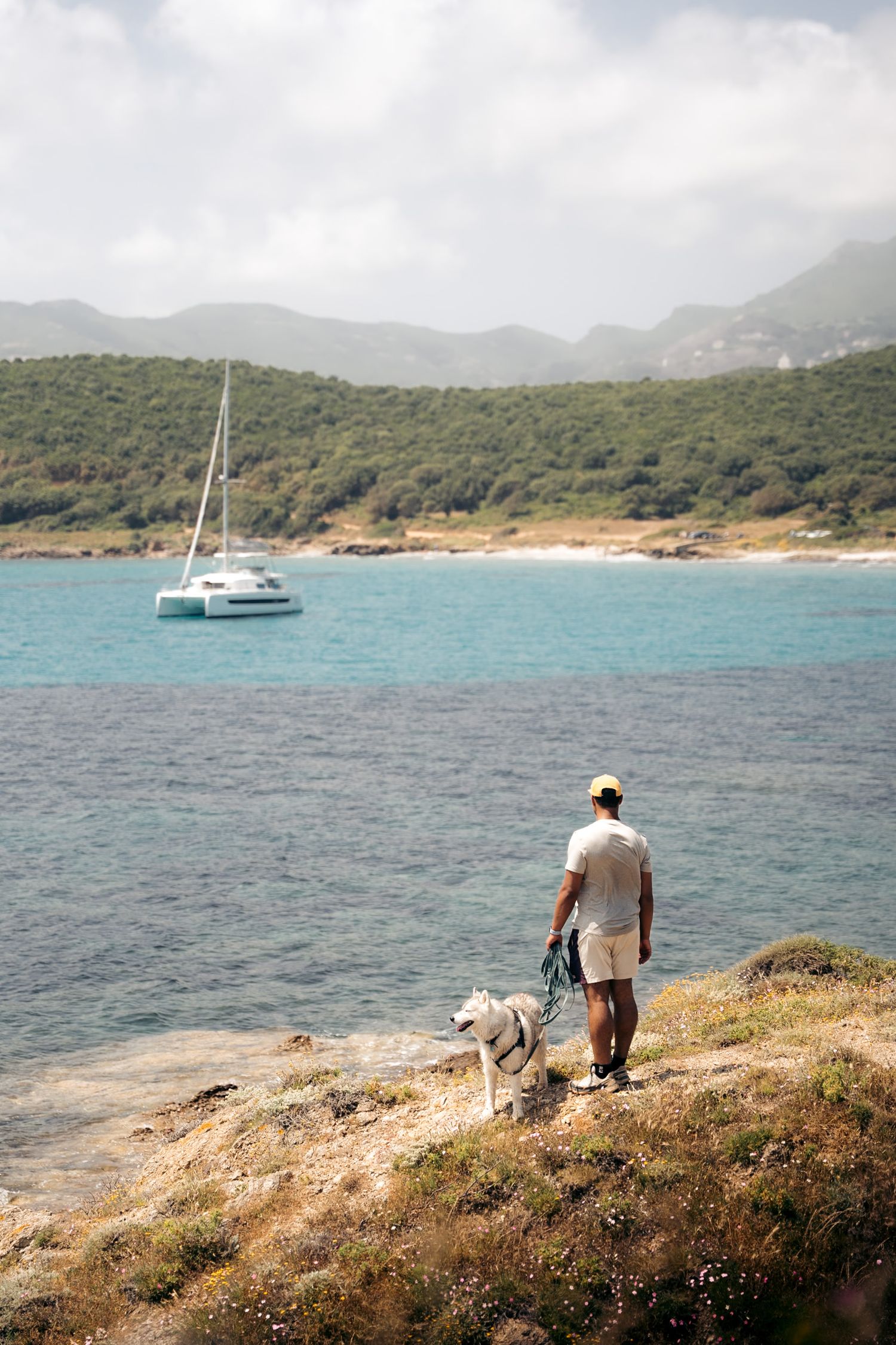 promeneur et son chien sur le sentier des douanier au cap corse avec un voilier dans la baie et montagnes en arrière-plan