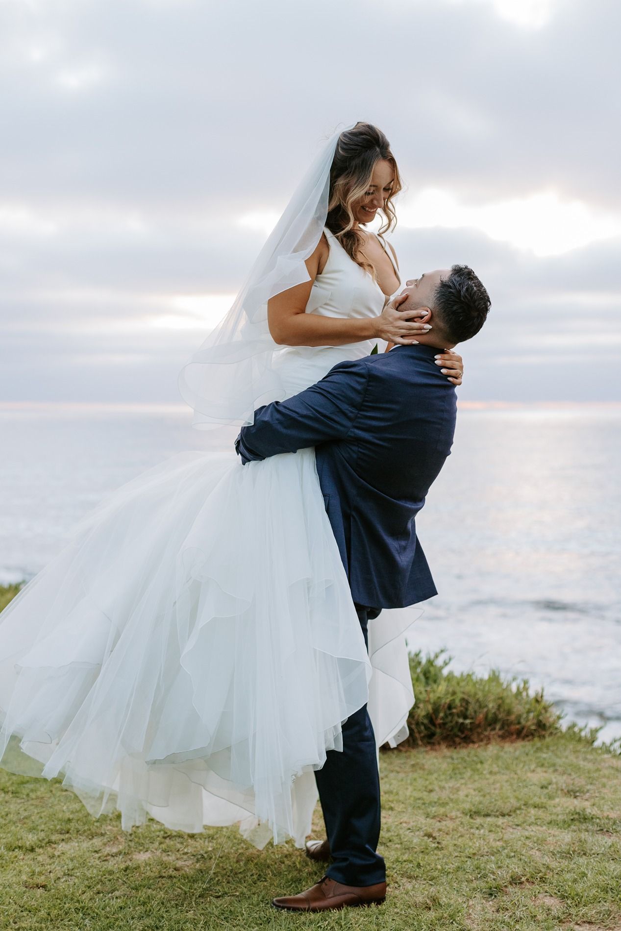 Groom in navy suit lifts bride in white tulle gown and veil by the ocean at sunset in La Jolla.
