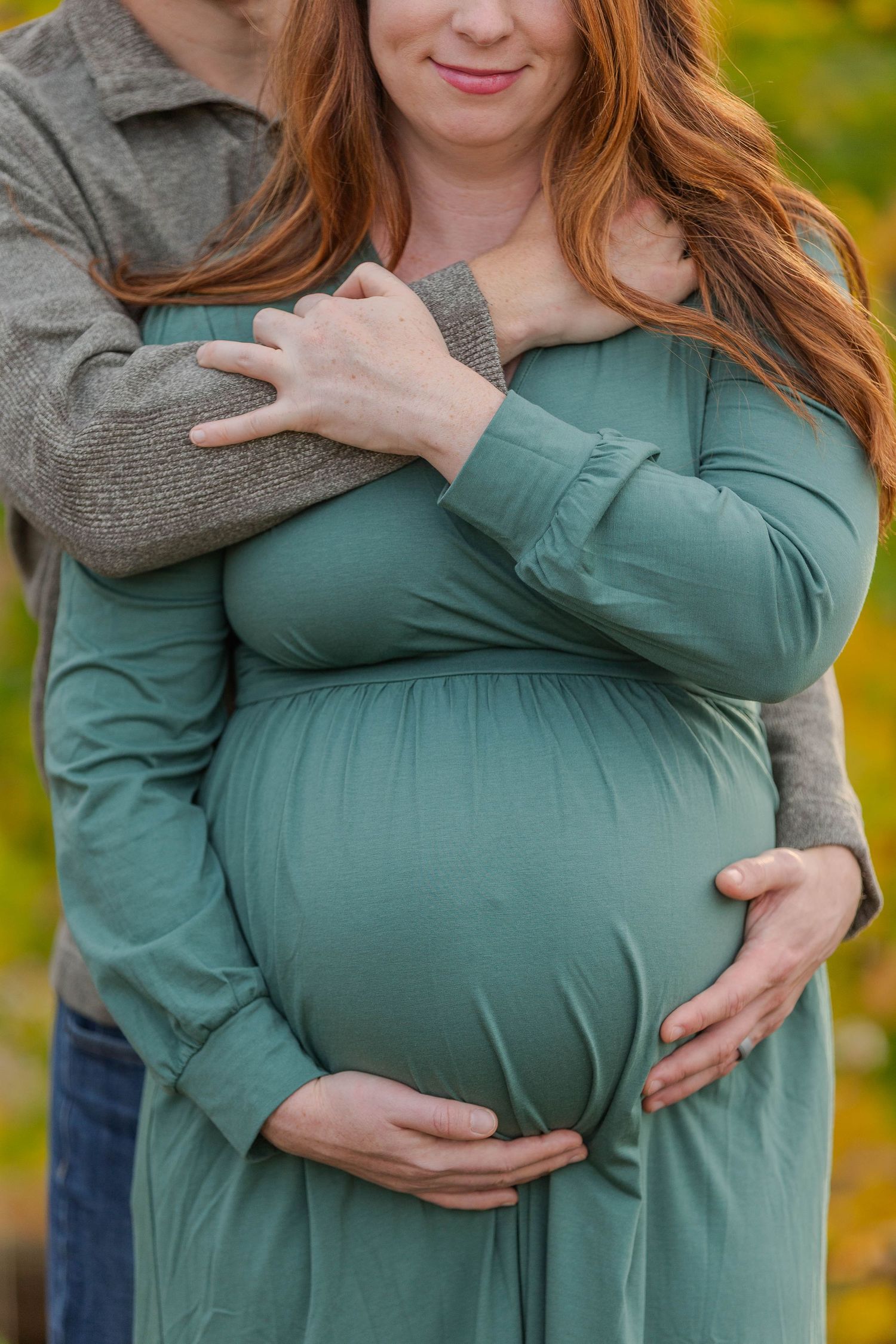 Pregnant woman in teal dress cradled by partner outdoors during maternity photo session.