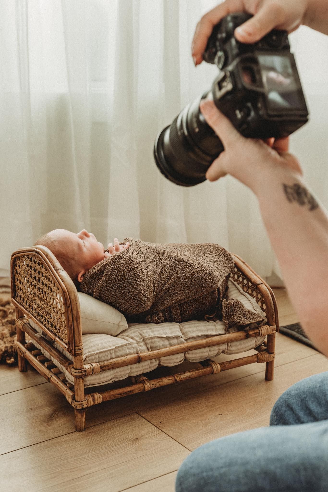 Newborn baby wrapped in brown knit swaddle on rattan mini bed during professional photography session.