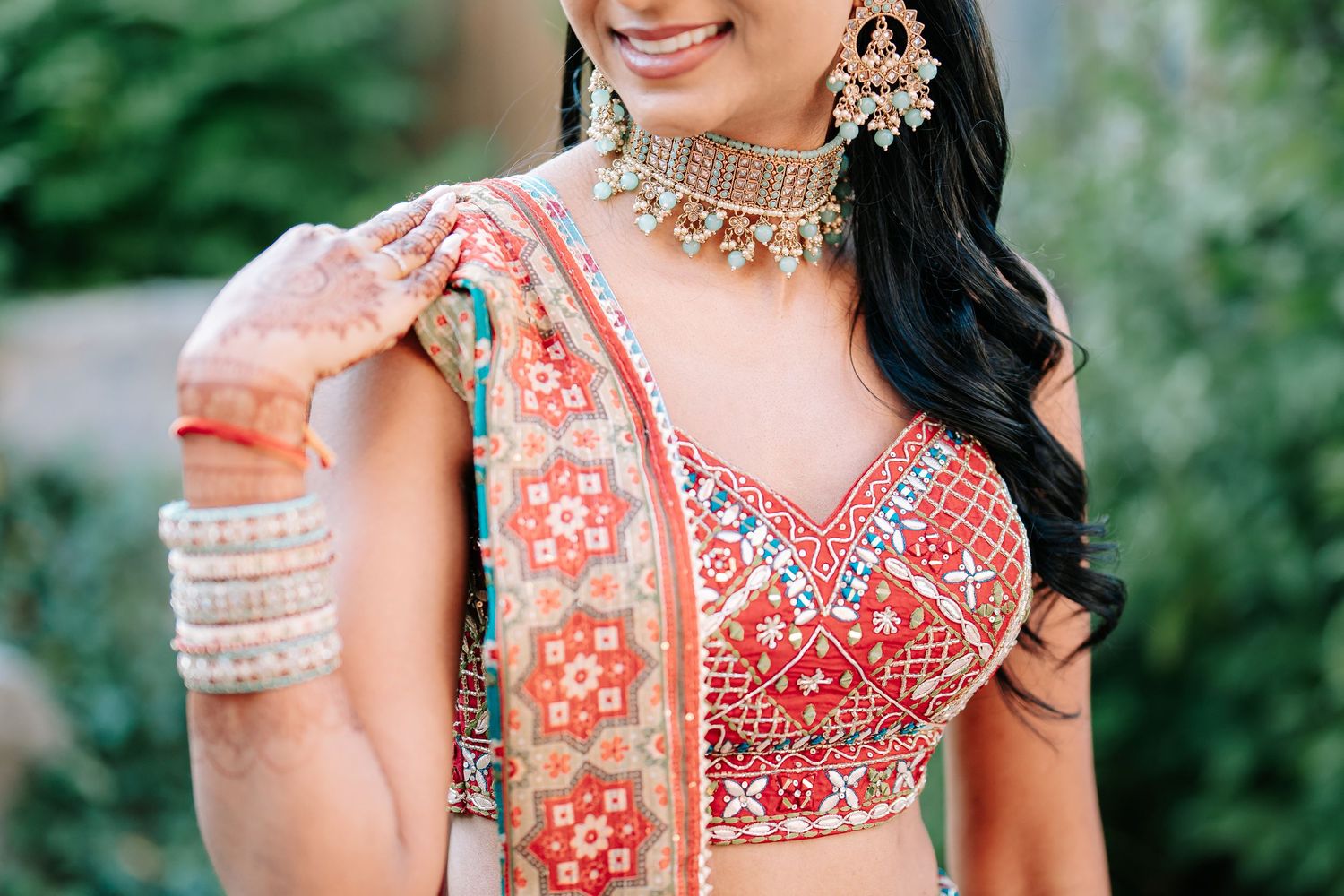 Bride in vibrant red embroidered lehenga with ornate choker necklace and mehndi henna on her hands.