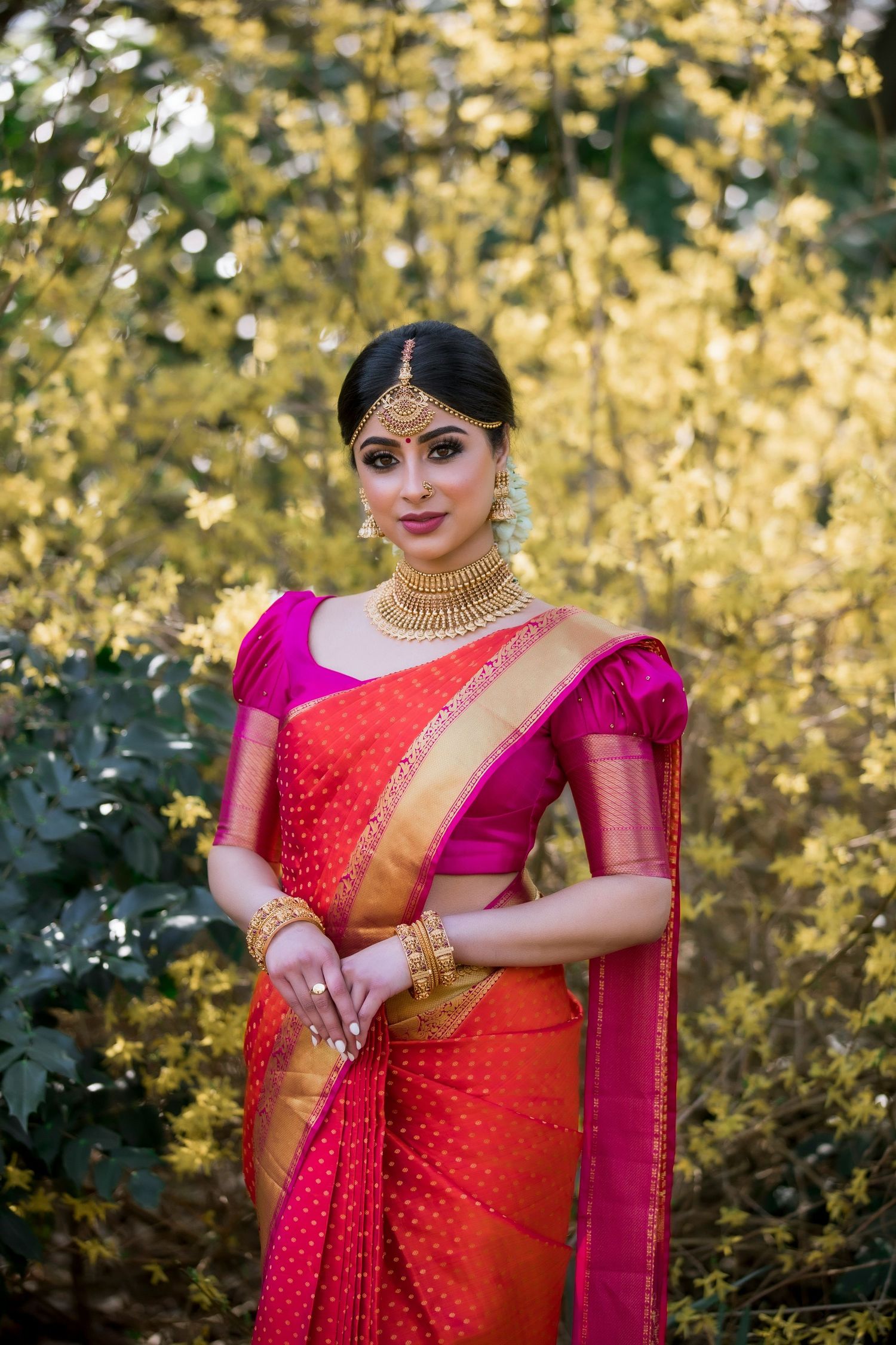 Hindu Bride in Austin, TX in red and gold saree with pink blouse and gold jewelry poses outdoors against lush green foliage.