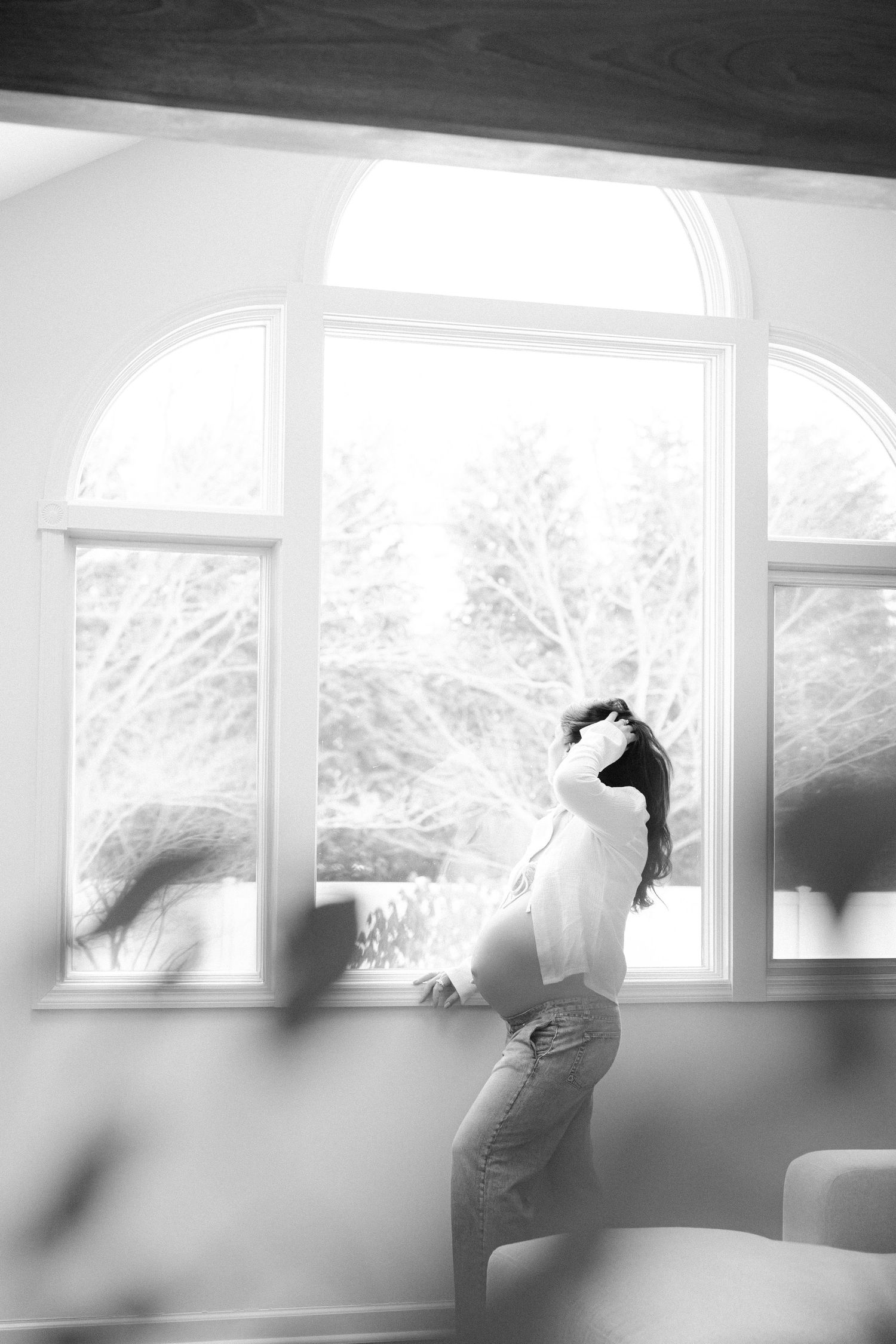 Black and white photo of a pregnant woman standing by large arched windows, cradling her baby bump in natural light.