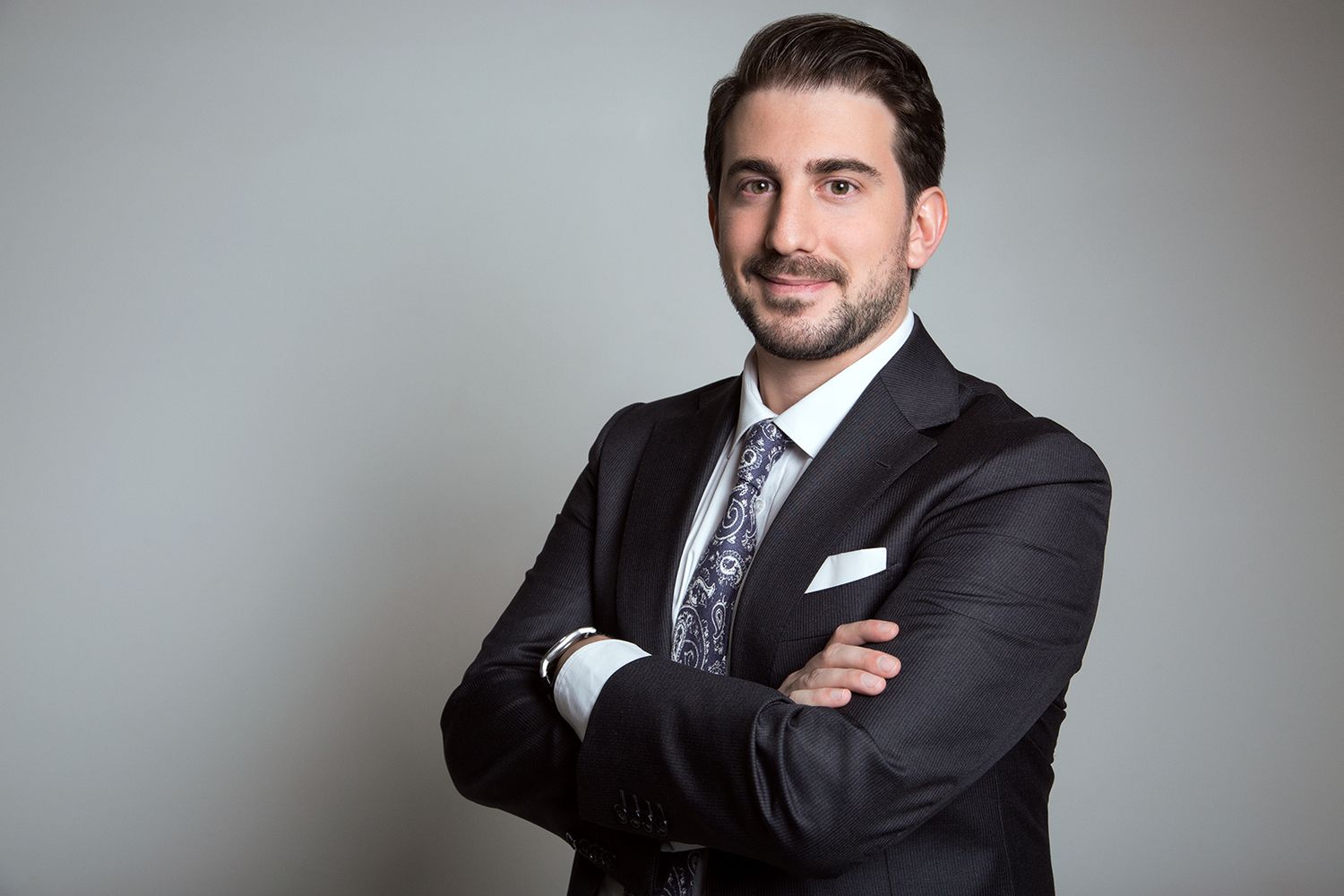 Confident businessman in dark suit with arms crossed posing against gray background.