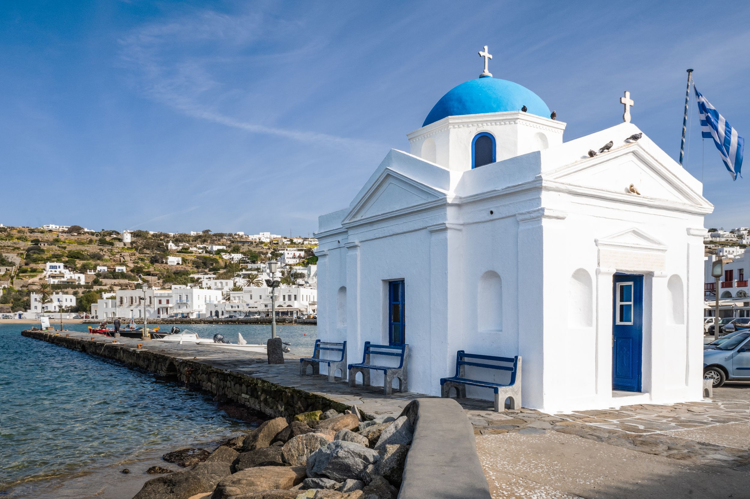 A series of white Greek Orthodox churches with blue domes and crosses line the Mediterranean coastline in Mykonos, Greece.