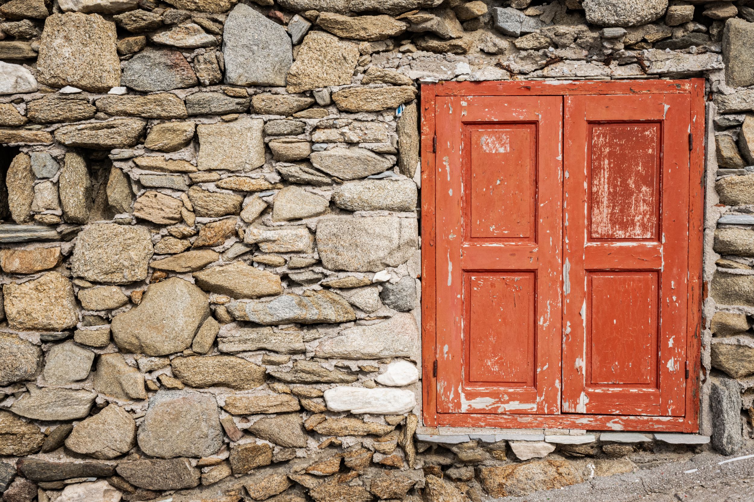 Row of rustic stone walls with red wooden shutters and doors along a Mediterranean building facade.