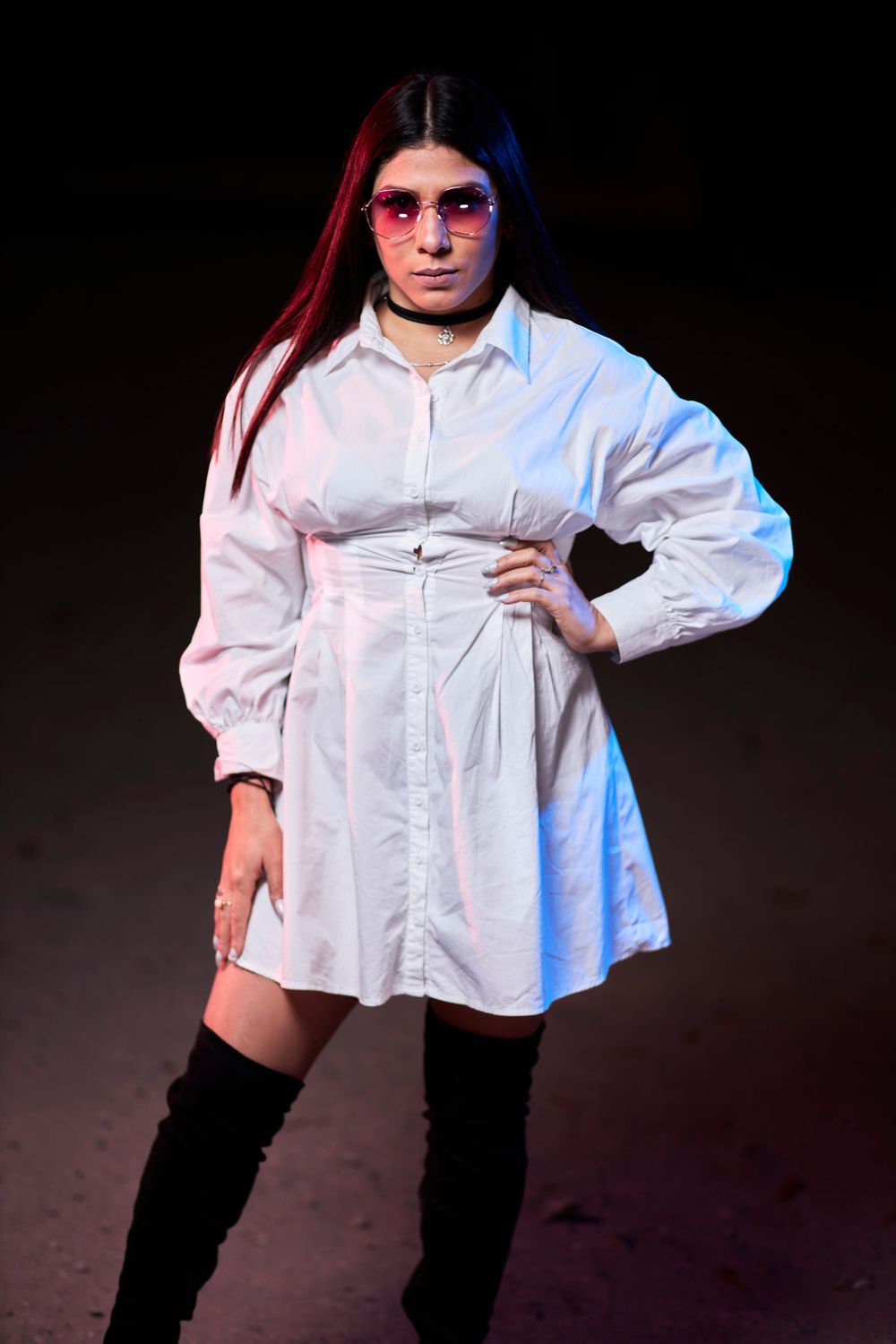 Fashion photo of model in white shirt dress and boots with dramatic red and blue lighting.