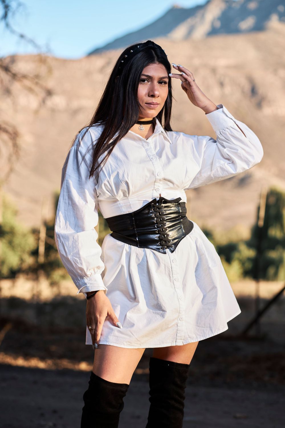 Fashion model in white shirt dress with black corset belt and thigh-high boots poses against desert mountains.