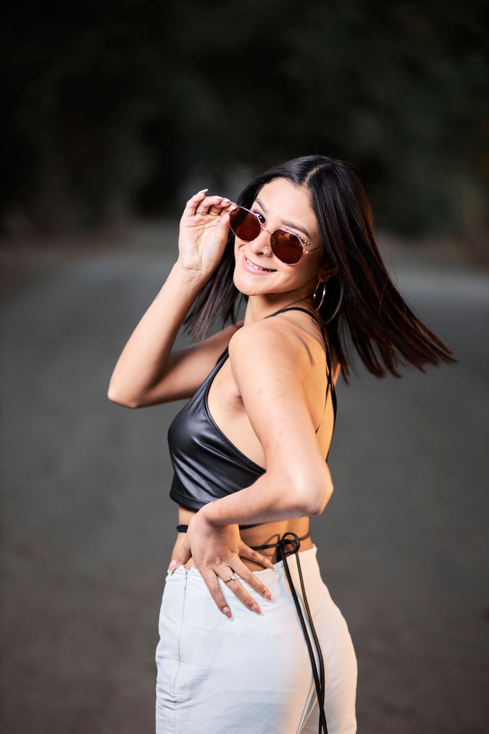 Model in leather crop top and white pants poses playfully with sunglasses on dirt road.