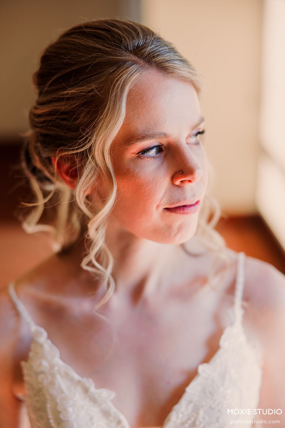 A bride in a white wedding dress with delicate lace details looks to the side in a soft, natural light setting.