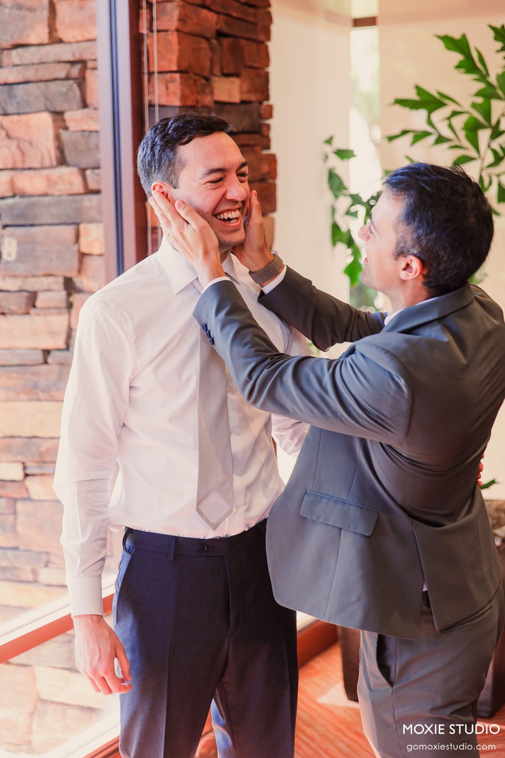 Two people in formal attire share a joyful moment in front of a brick wall with greenery.