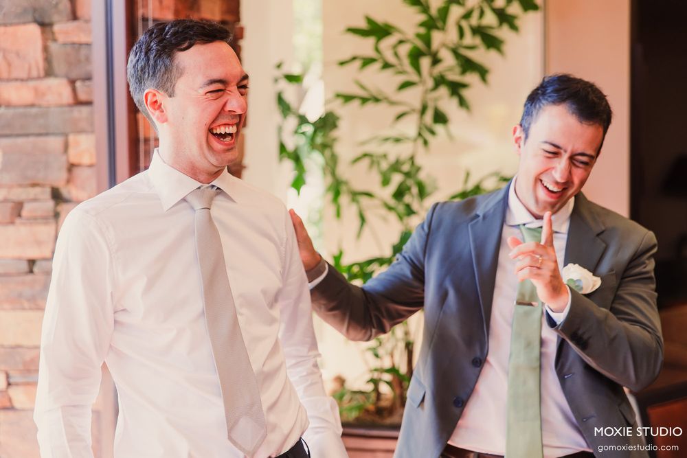 Two men in formal attire share a candid laughing moment at an indoor venue with brick walls and greenery.