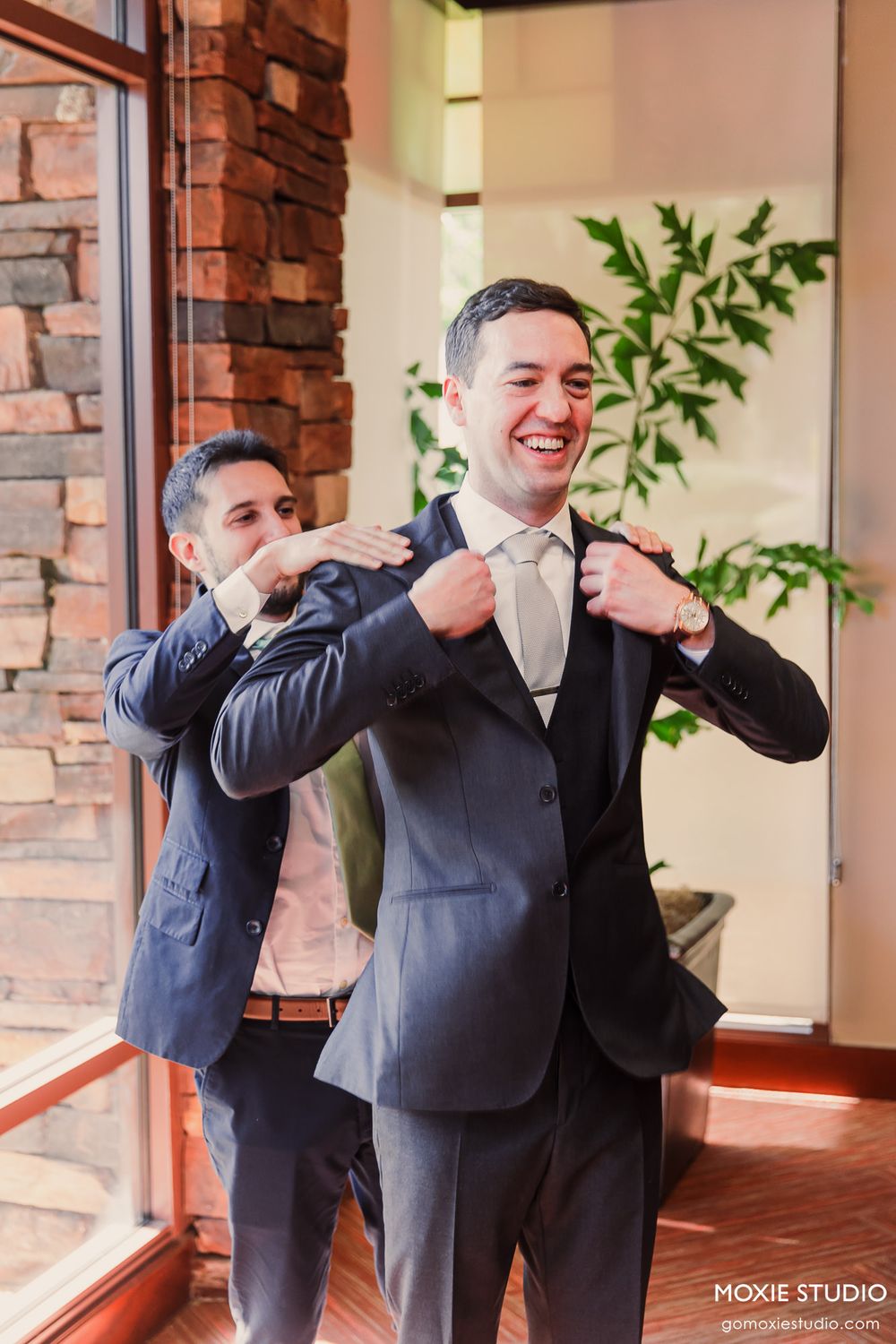 Groomsmen in dark suits sharing a fun candid moment dancing and posing in a brick-walled indoor venue.