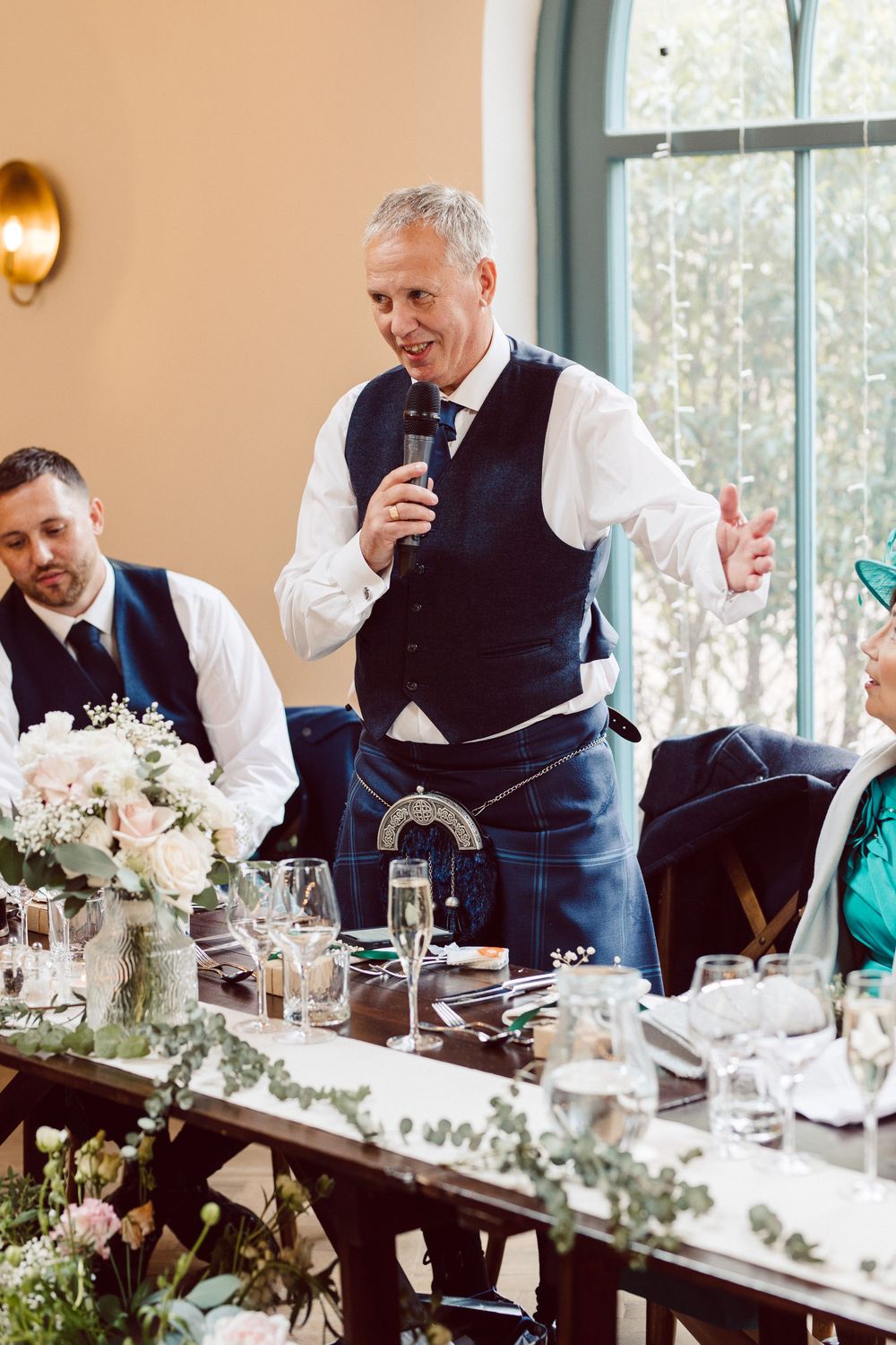 Wedding guests in formal attire seated at an elegant table with floral centerpieces and glassware in a sunlit room.