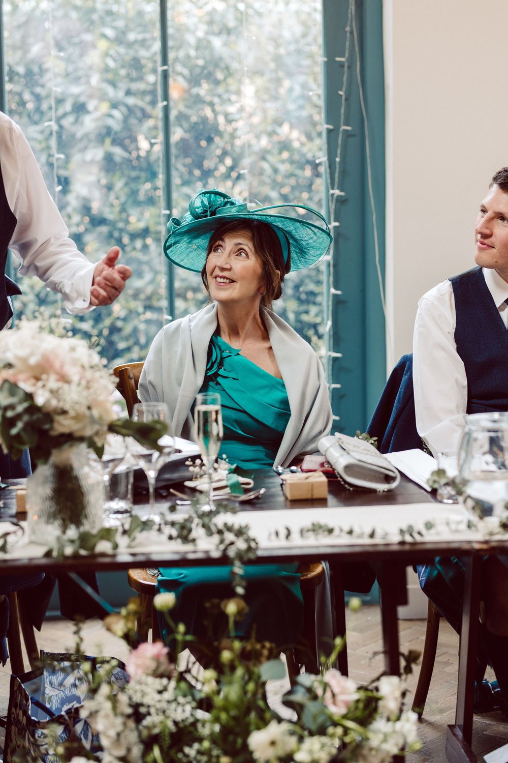 Wedding party guests seated at long table decorated with white floral arrangements laugh and celebrate during reception.