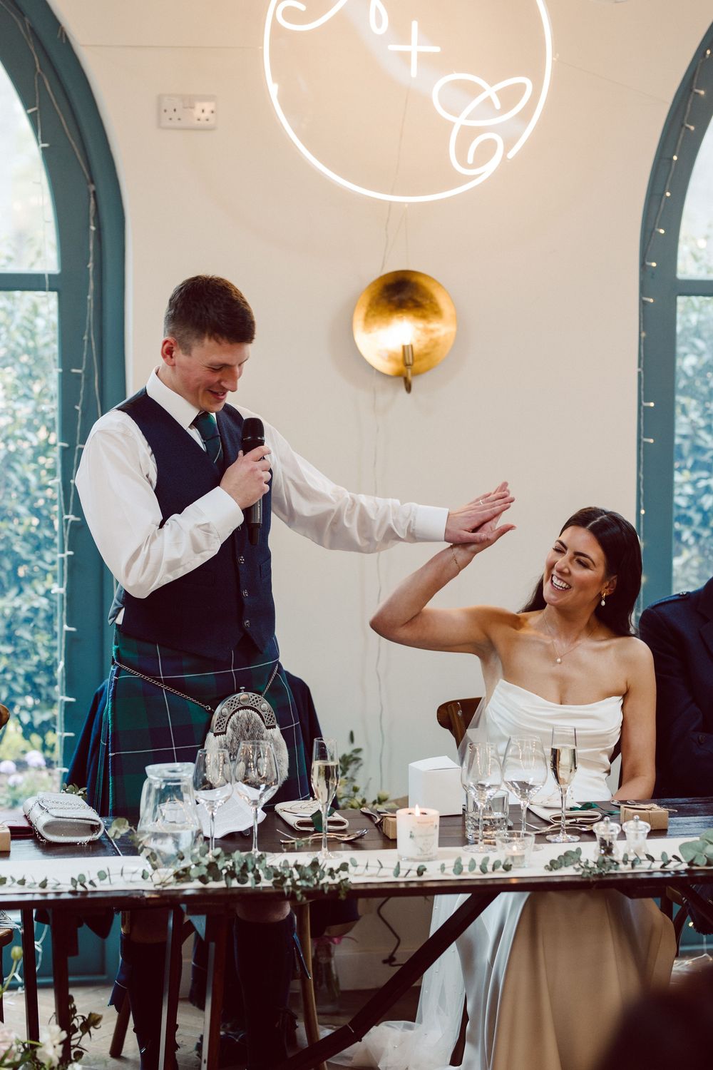 Wedding reception scene at a venue with arched windows showing a couple and guests seated at an elegant dining table.