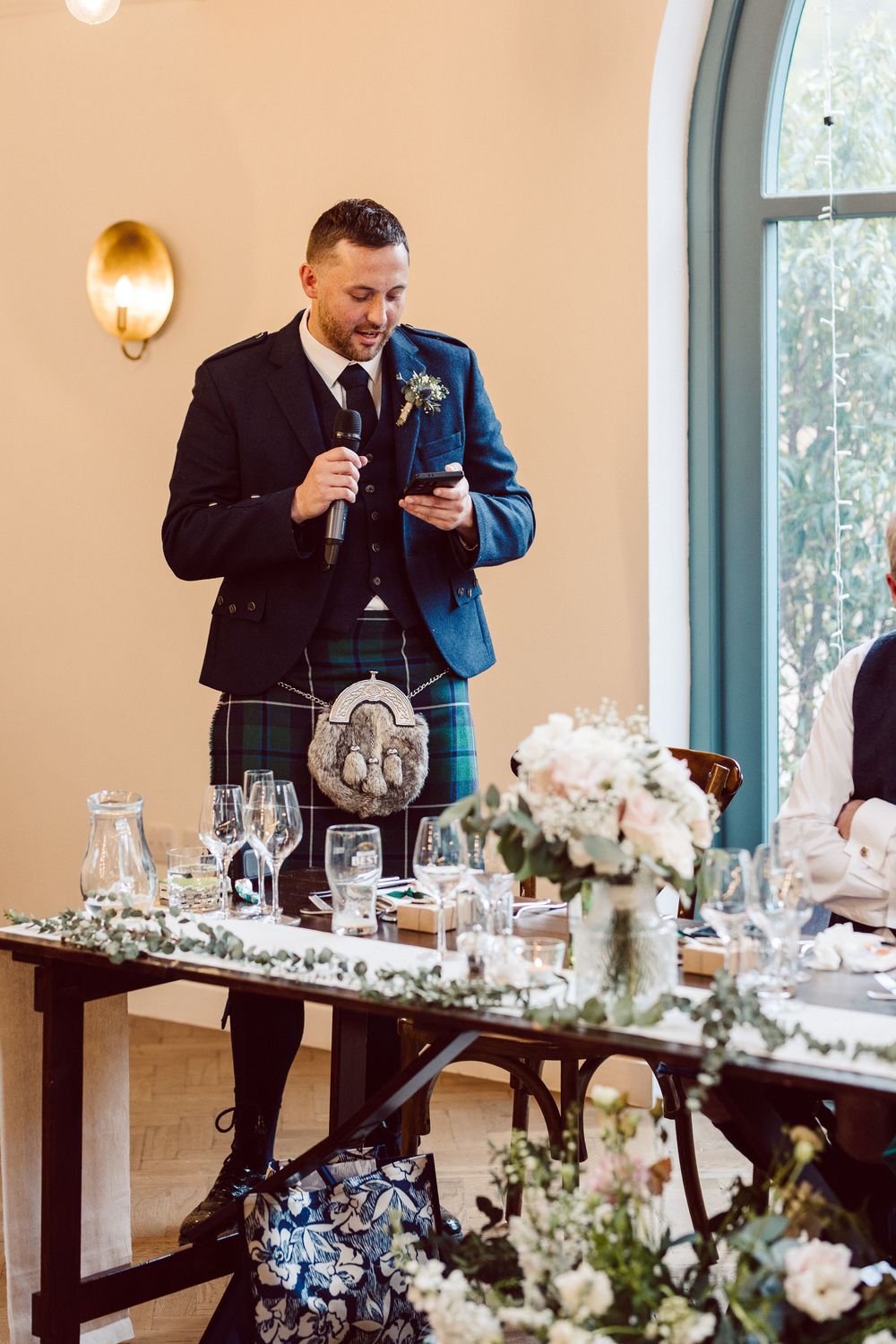 A person in traditional Scottish kilt and formal jacket gives a speech at an elegant wedding reception table with floral decor.