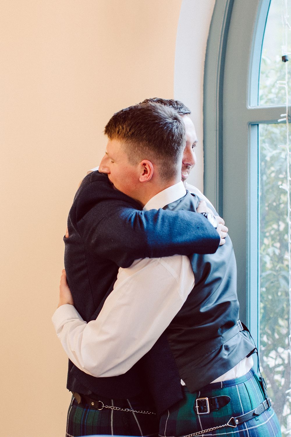 Two people wearing kilts embrace in an emotional moment by a window with natural light streaming in.
