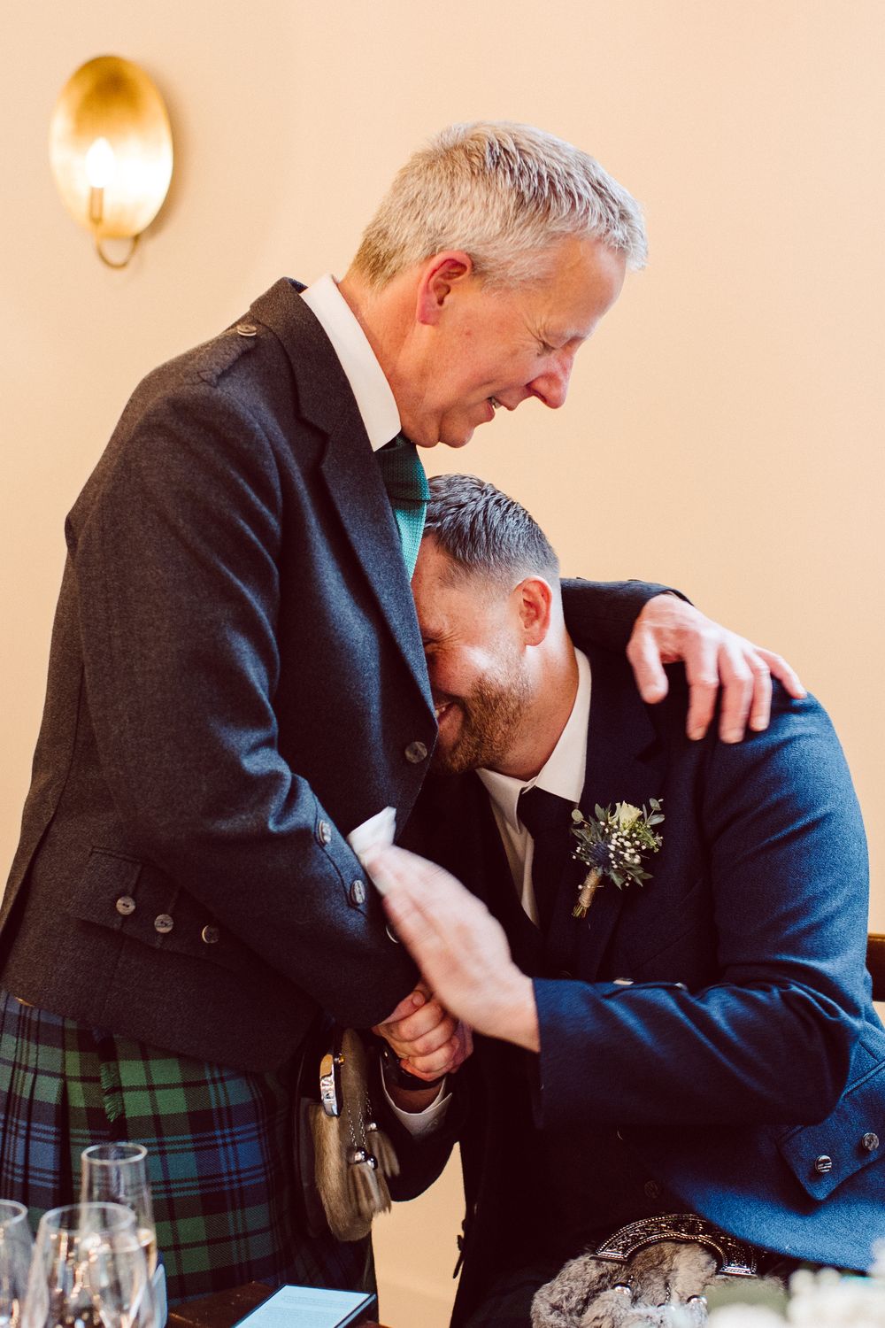 Two men in formal attire and kilts share an emotional embrace at a table during what appears to be a wedding reception.