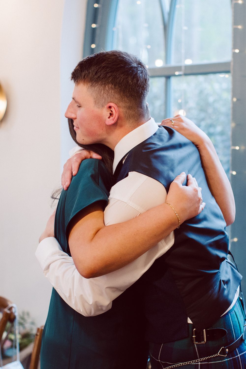 Two people share an emotional embrace during a wedding celebration with twinkling lights in the background.