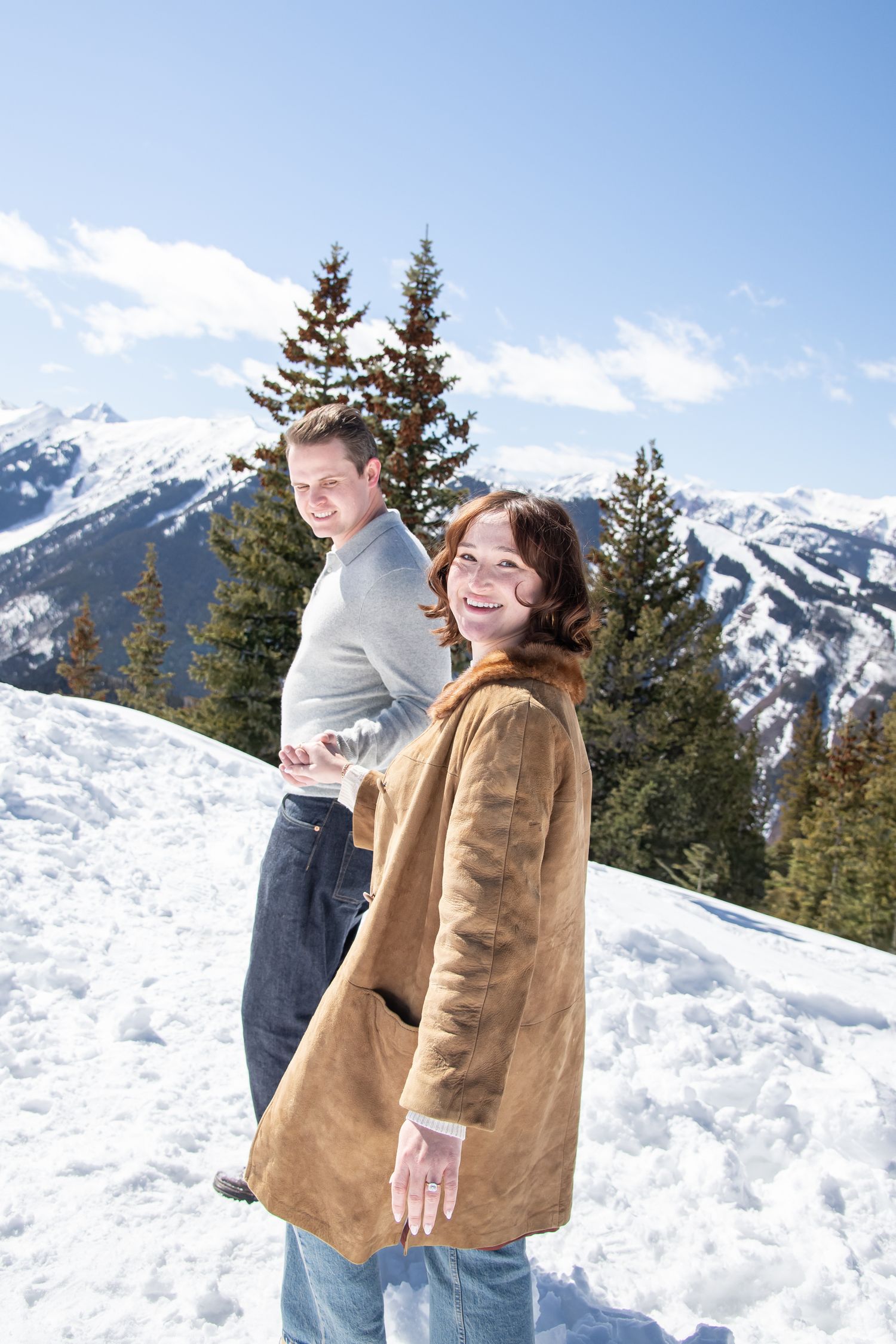 A couple shares a playful moment in the snow with snow-capped mountains and evergreen trees in the background for Aspen Colorado photos.