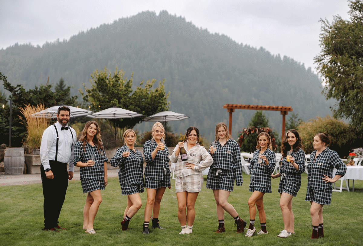 Wedding party in matching plaid shirts and denim performs synchronized dance moves on a grassy lawn with mountains behind.