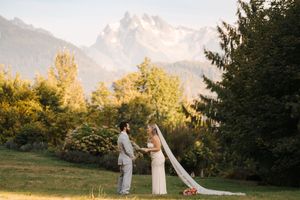 A couple in wedding attire poses in a scenic mountain meadow at sunset with evergreen trees in the background.