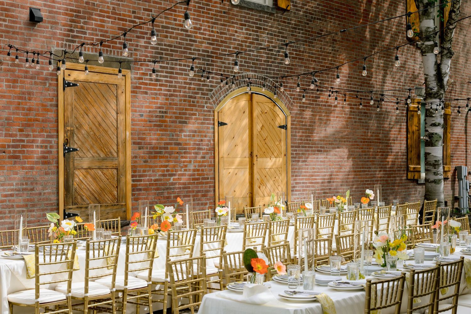 Long banquet table with white linens and wooden chairs set up in a rustic brick venue with string lights overhead.