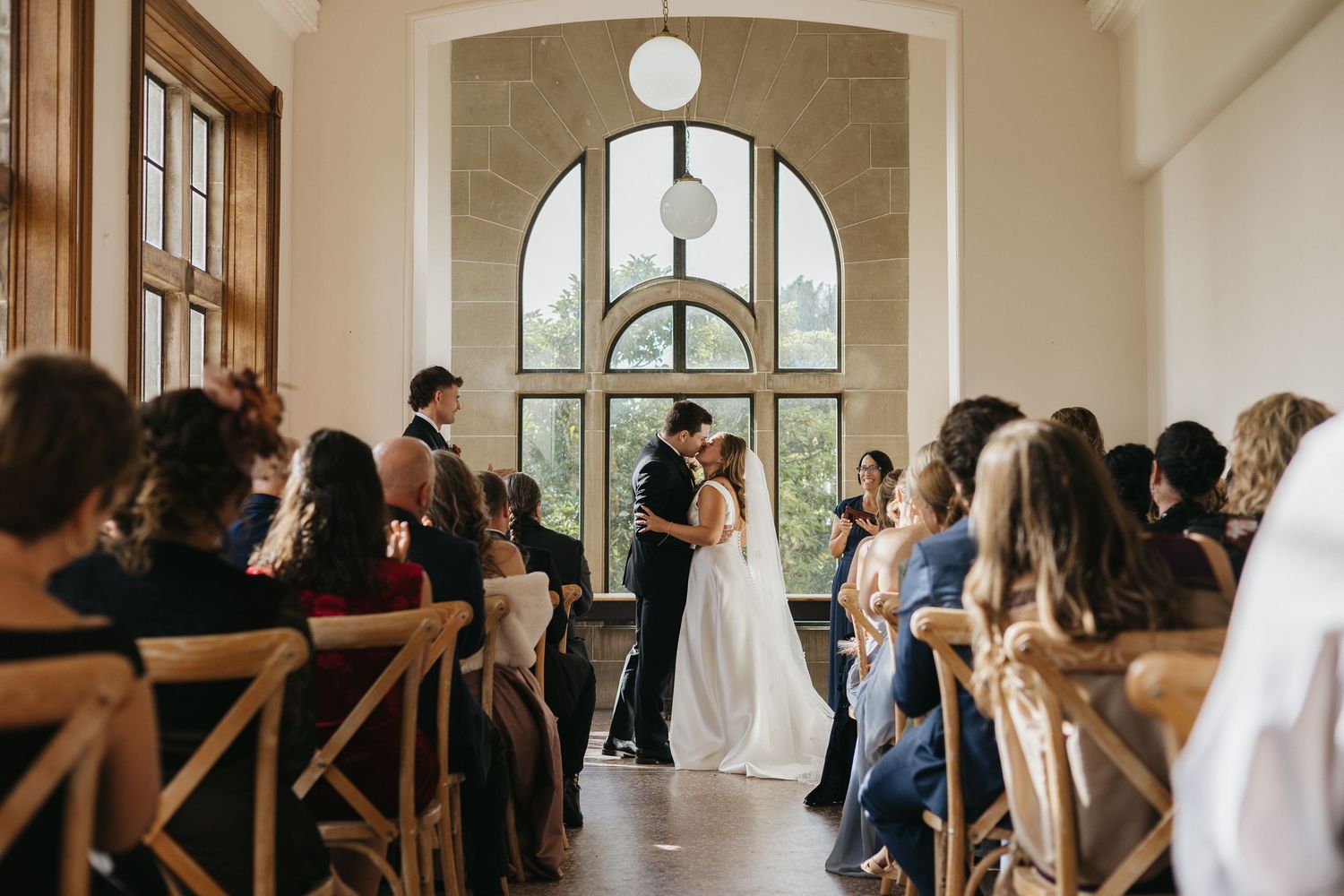 Panoramic view of a wedding ceremony in a bright white room with large windows and guests seated in rows.