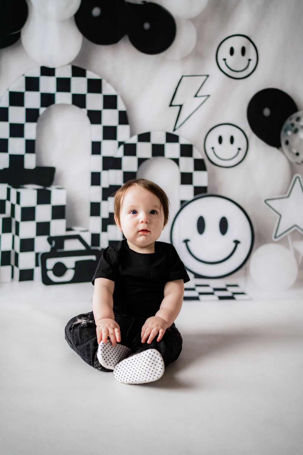 Baby in black outfit sits before black and white checkered decorations and smiley face balloons.