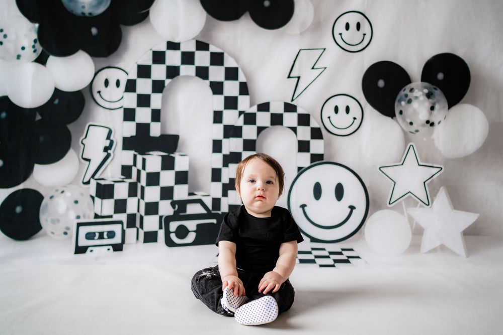 Baby in black outfit sits before black and white smiley face balloon backdrop with checkered decor.