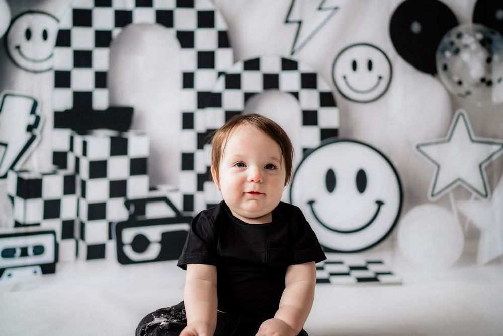Baby in black onesie sits before black and white checkered backdrop with smiley face balloons.