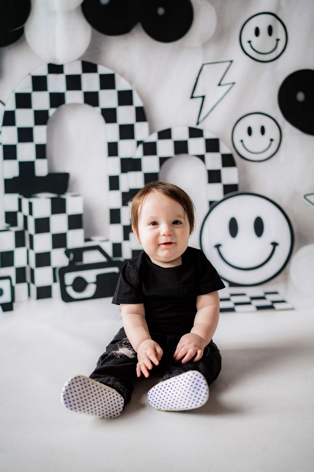 Smiling baby in black outfit sits in front of black and white smiley face birthday party backdrop.