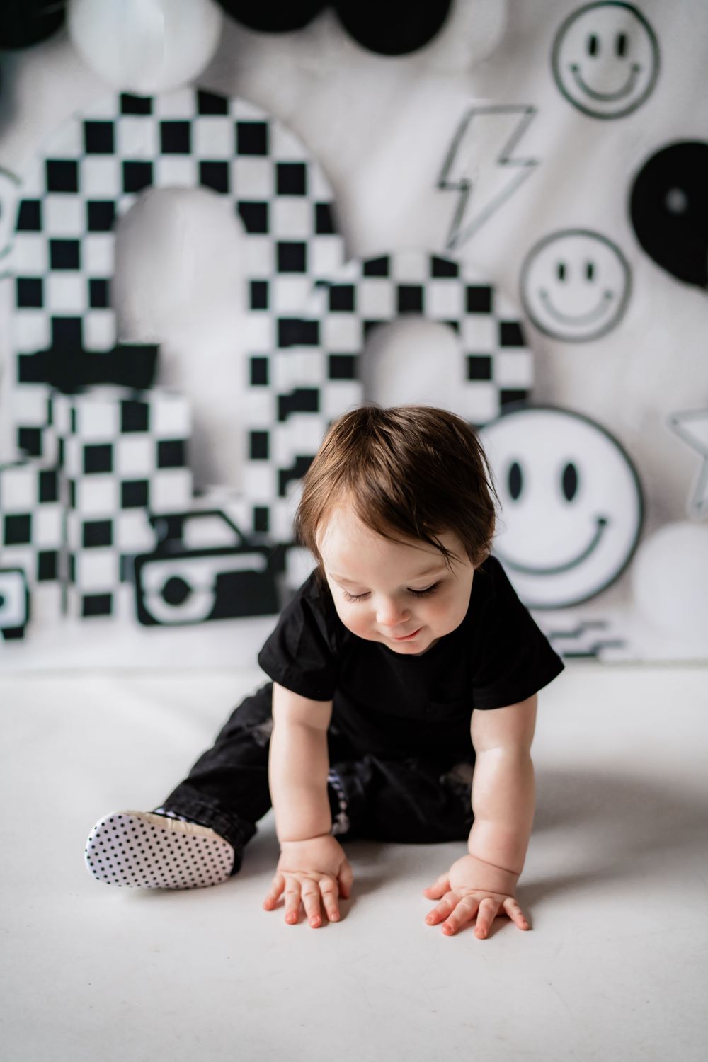 A baby in black outfit crawls on white floor with black and white smiley face birthday party decorations behind them.