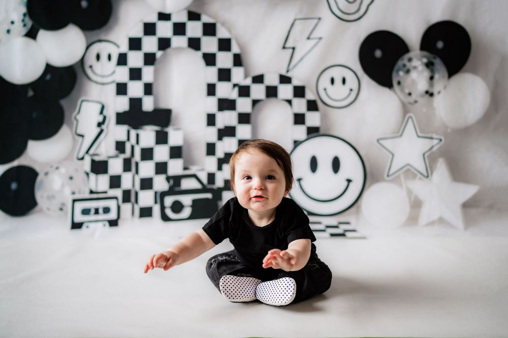 Baby in black outfit sits cross-legged before black and white smiley face party backdrop.