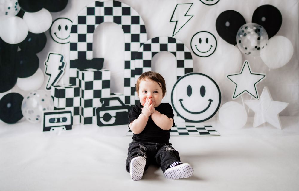 Baby in black outfit sits before checkered backdrop with smiley face balloons at a first birthday photo shoot.