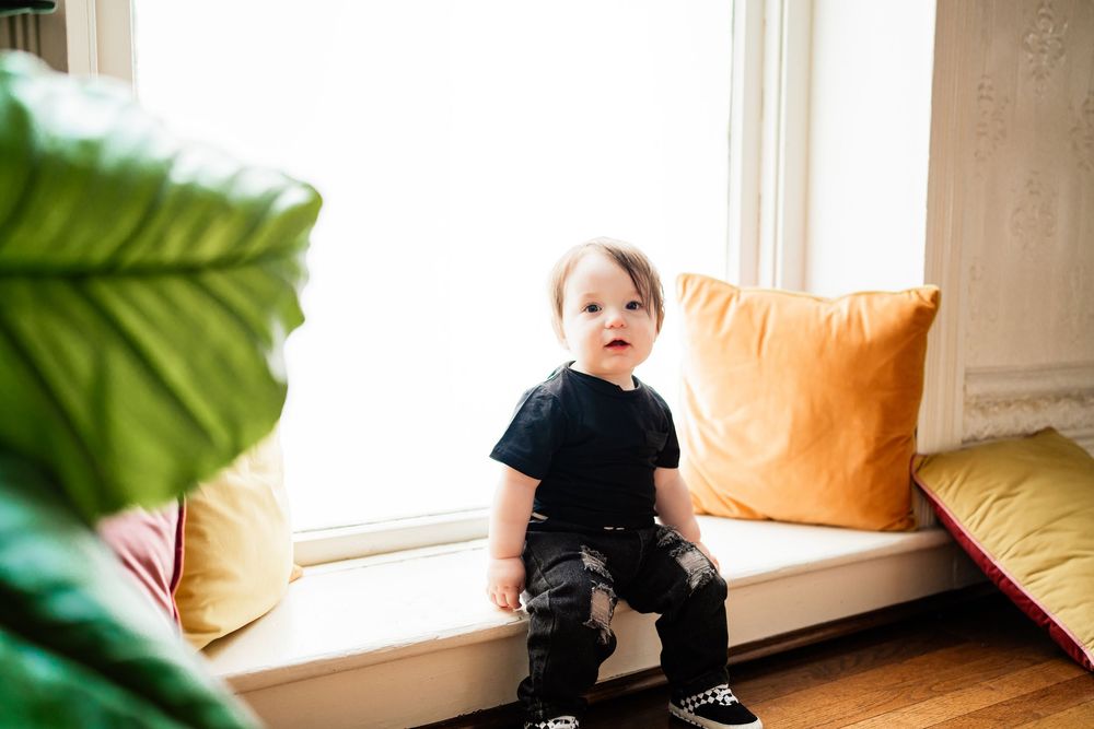 A curious toddler in a black outfit stands on a window seat surrounded by colorful cushions and tropical leaves.
